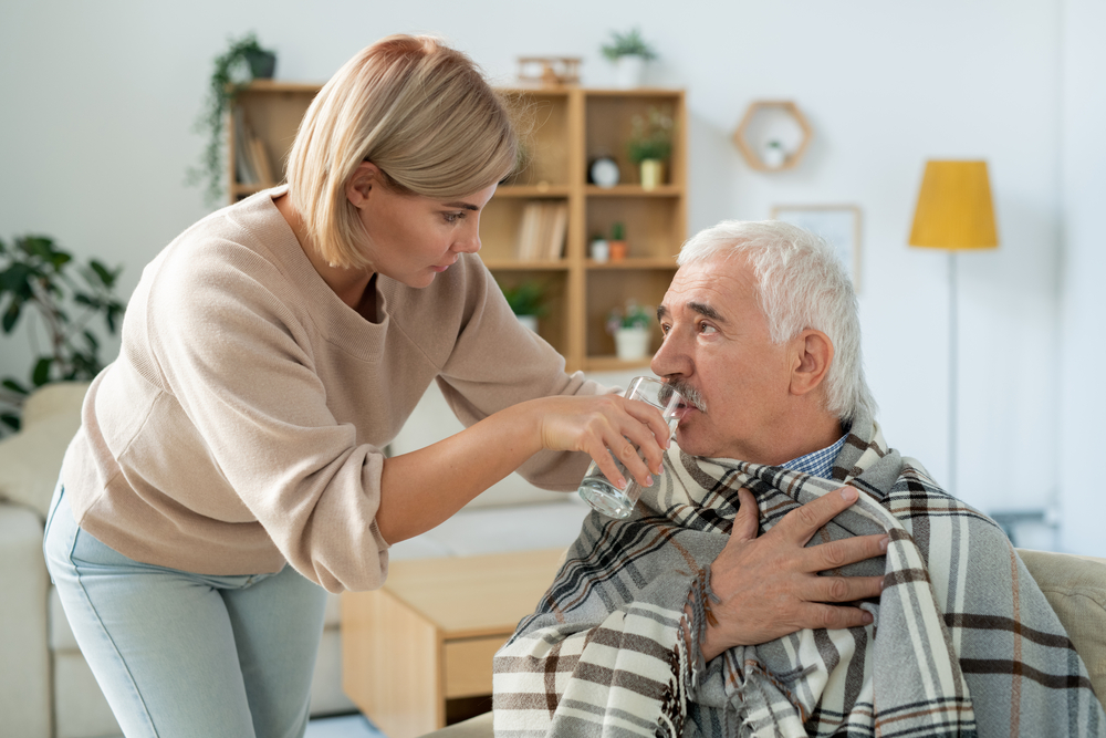 Careful young woman taking care of her sick senior father wrapped in plaid while giving him glass of water